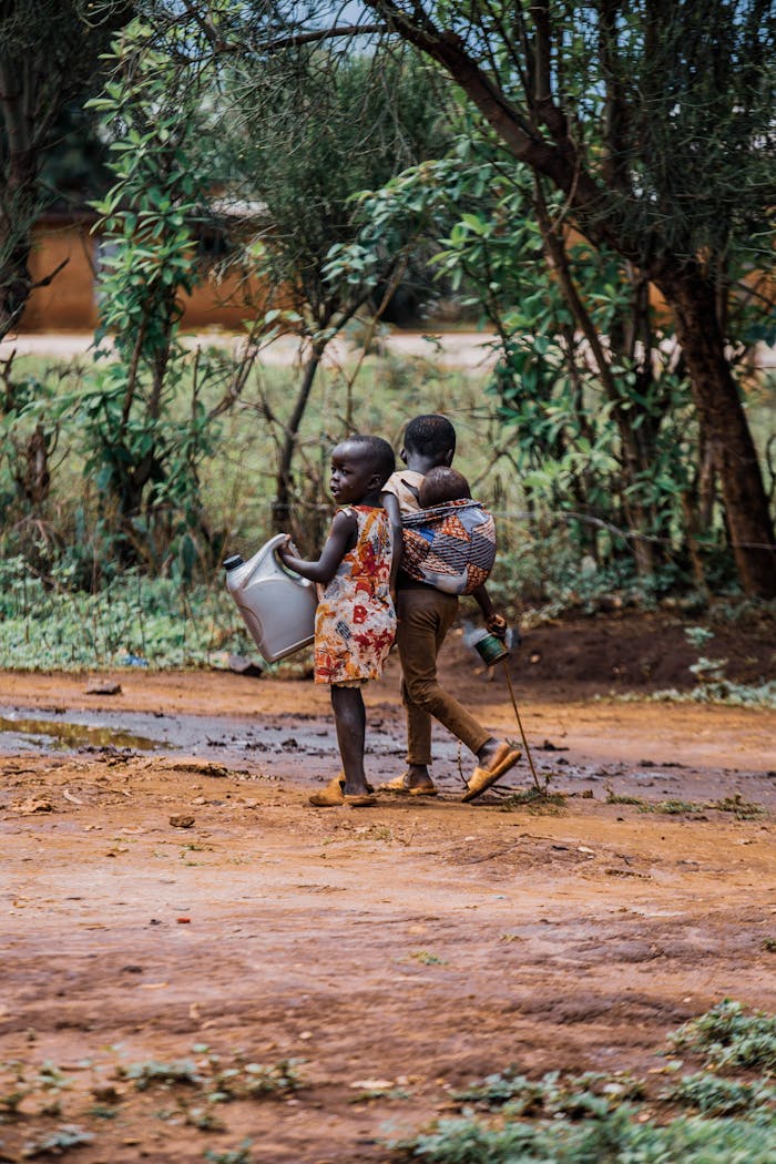 Two children carry water and a sibling along a rural muddy path.